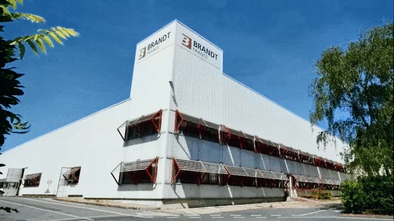 Vue extérieure d'un bâtiment industriel de la marque Brandt, avec un ciel bleu clair et de la verdure en arrière-plan.