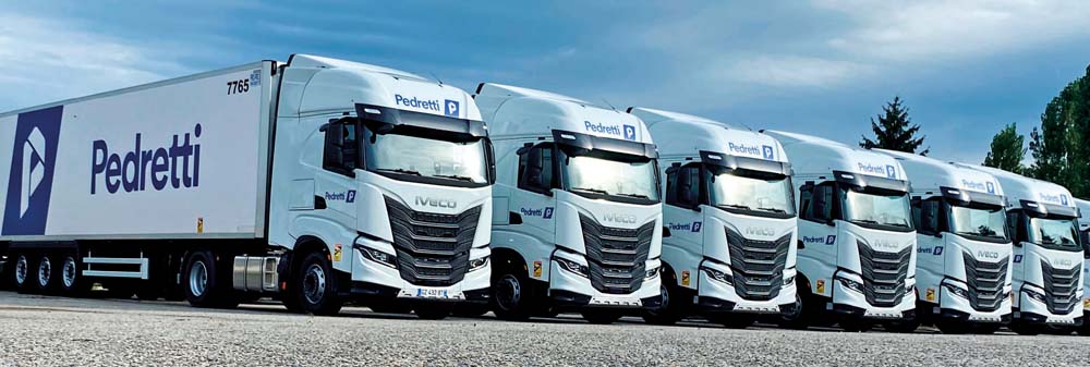 Une flotte de camions IVECO blancs avec la marque Pedretti, garés en ligne sur un terrain dégagé, sous un ciel nuageux.